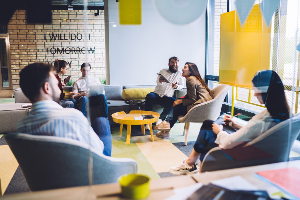 Group of joyful and cheerful coworkers sitting together in bright creative office and laughing while discussing launch of new project of company