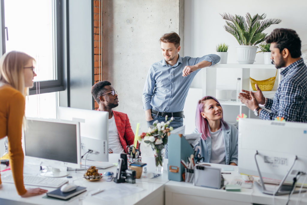 A group of young employees jovially collaborating at their work desks during a busy morning at the office.